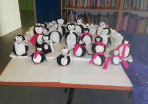 Na zdjęciu widać stojące na stole pingwiny, wykonane przez uczniów klasy 2b. Za stolikiem z pingwinami znajduje się regał z książkami.